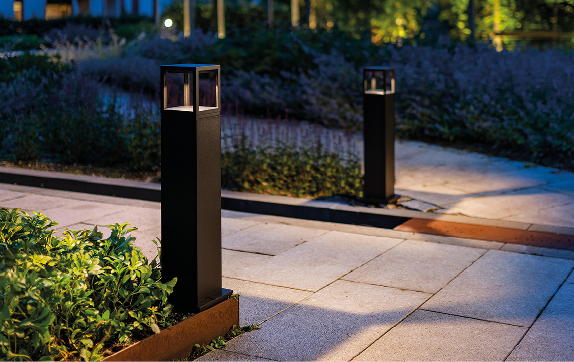 Bildet viser 2 stykk av pullerten Fagernes Bollard, som lyser opp på hver sin side av en steinbelagt gangvei. Foto.