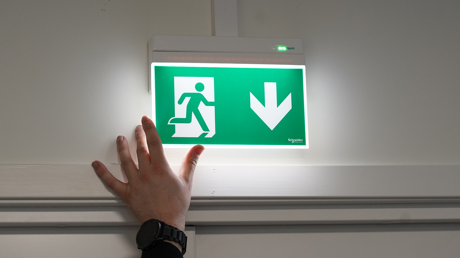 Bildet viser hånda til en elektriker som justerer et nødlys fra Schneider, i Exiway-serien. Foto.