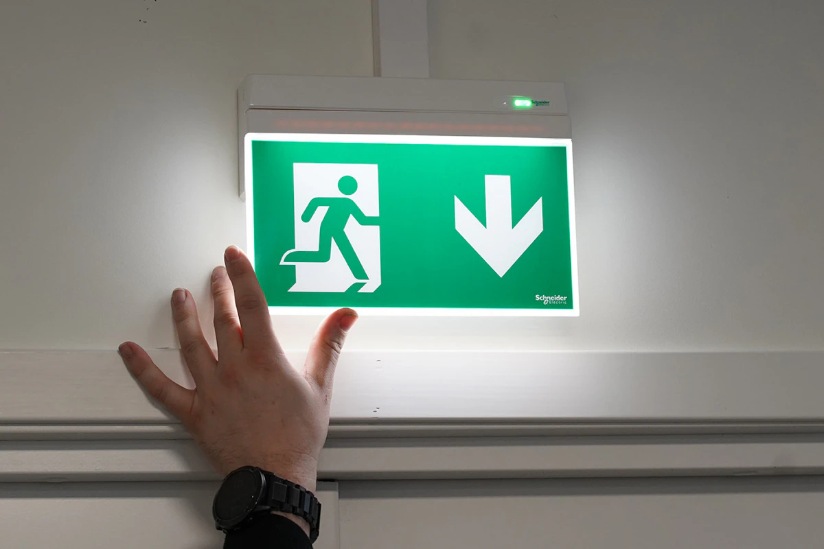 Bildet viser hånda til en elektriker som justerer et nødlys fra Schneider, i Exiway-serien. Foto.