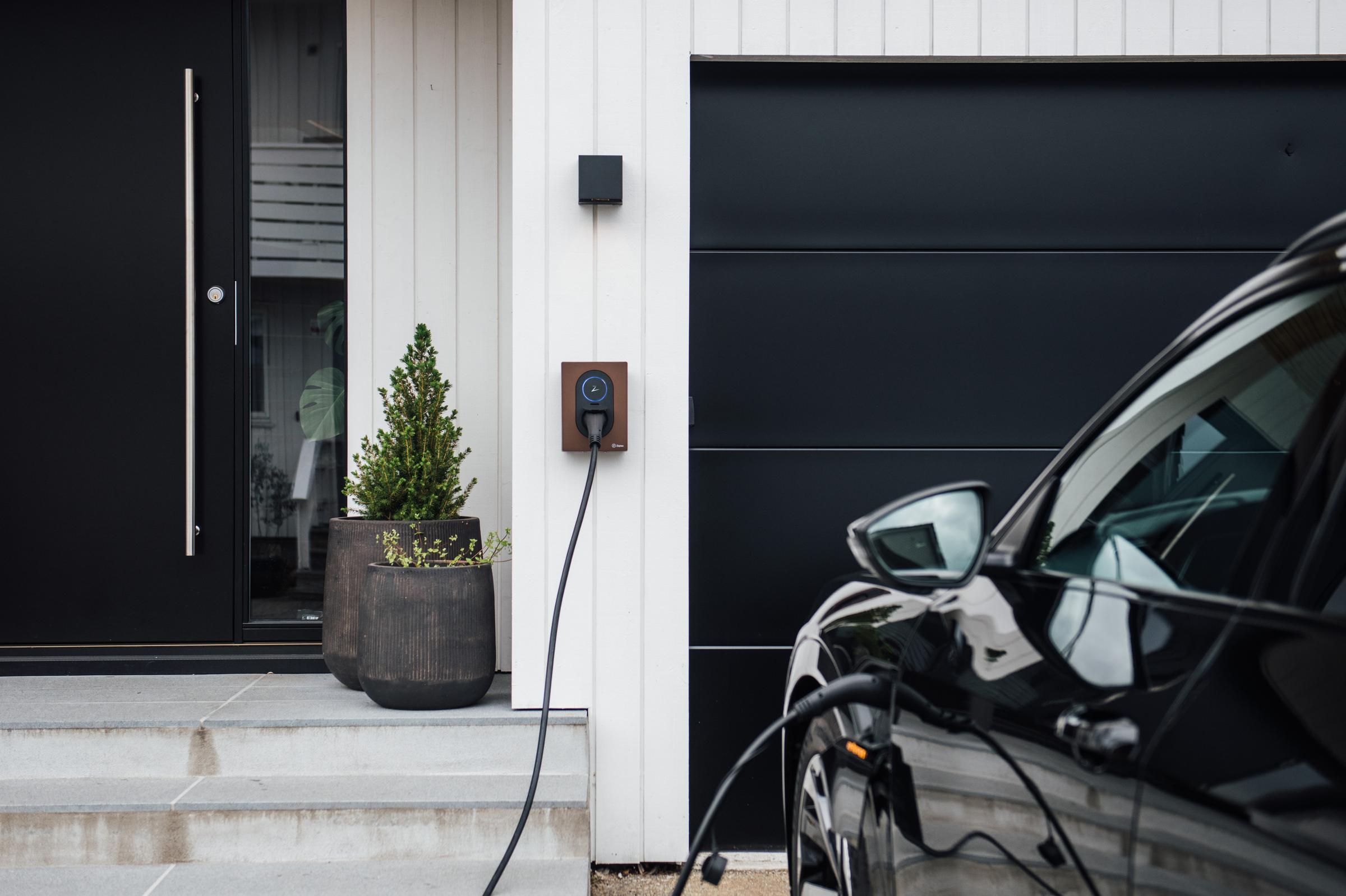 Zaptec GO elbillader i sort på hvit vegg koblet til en sort bil. Bilde