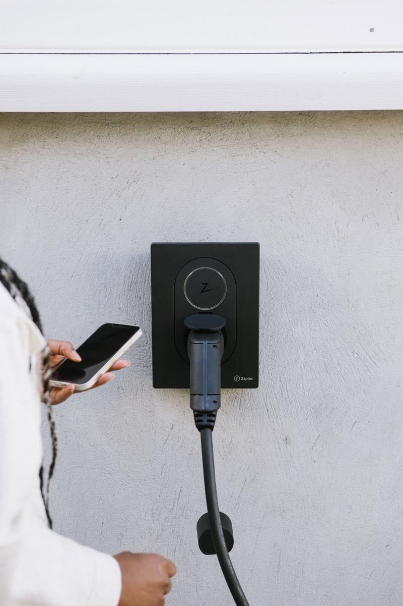 Zaptec GO elbillader i fargen sort montert på hvit vegg med en dame som styrer laderen med en app. Bilde