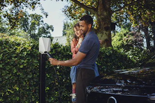 Laderen Easee Charge Up i fargen hvit, montert på en stolpe ved en hekk, og en far og datter som kobler bilen til laderen. Foto.