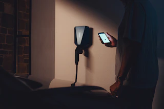 Laderen Easee Charge Up i fargen svart, montert på en vegg, og et menneske som er på mobilen sin. Foto.